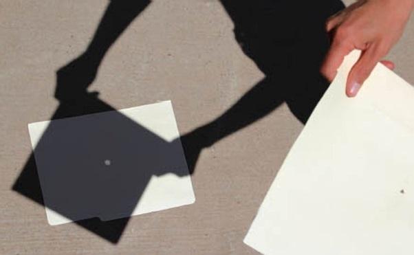 A piece of paper with a small hole cut in it (known as a pinhole camera) is held above the ground. A small image of the sun can be seen on a piece of paper on the sidewalk through the pinhole camera.